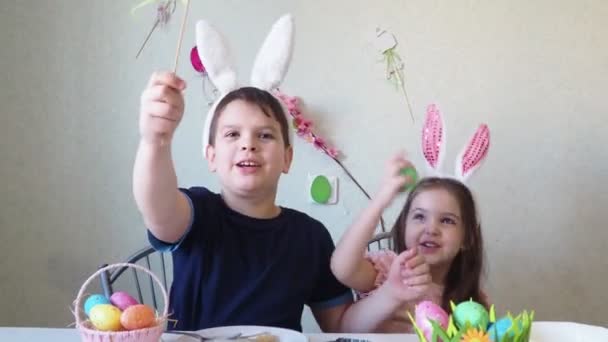 Jongen en meisje bereiden koekjes voor Pasen, lachen. Plezier hebben op de paaseieren jacht. Kind en meisje met konijnenoren en eieren aan. kleurrijke eieren. — Stockvideo