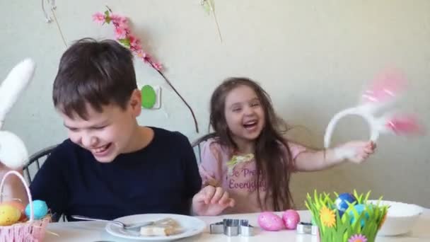 Chico y chica están preparando galletas para la Pascua, riendo. Divertirse en la búsqueda de huevos de Pascua. Niño y niña usando orejas de conejo y pintando huevos. huevos coloridos. — Vídeo de stock