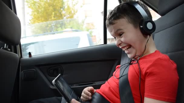 Joyeux petit garçon dans la voiture assis attaché avec une ceinture, siège et jouer avec tablette PC portant un casque, regarder des dessins animés, communiquer. voyage amusant. — Video