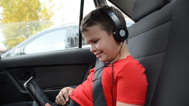 Šťastný chlapeček v autě sedí připoutaný opaskem, sedadlo a hraje si s tabletovým počítačem ve sluchátkách, sleduje kreslené seriály, komunikuje. zábavný výlet. — Stock video