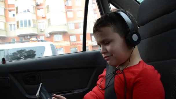 Szczęśliwy chłopiec w samochodzie siedzi zapinany paskiem, siedzeniem i bawi się tabletem nosząc słuchawki, oglądając kreskówki, komunikując się. wycieczka zabawy. — Wideo stockowe