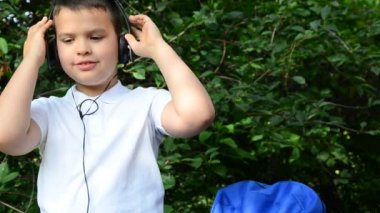 Parkta evrak çantasının yanında beyaz gömlekli bir çocuk kulaklıkla müzik çalıyor. Parktaki çimlerin üzerinde oturup müzik dersi dinleyen öğrenci.