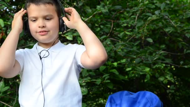 Een schooljongen in een wit shirt naast een aktetas in het park speelt muziek op een koptelefoon. Studenten leren luisteren audio tutorials in een park zitten op het gras in een park — Stockvideo