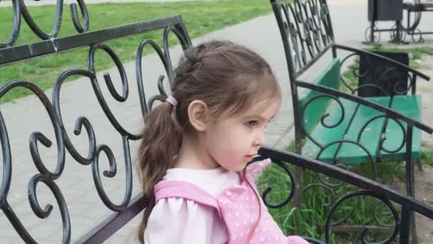 Chica en un banco del parque comiendo una dona con glaseado amarillo. infancia feliz, recreación al aire libre, paseos. Niño alegre sentado en el banco del parque — Vídeo de stock