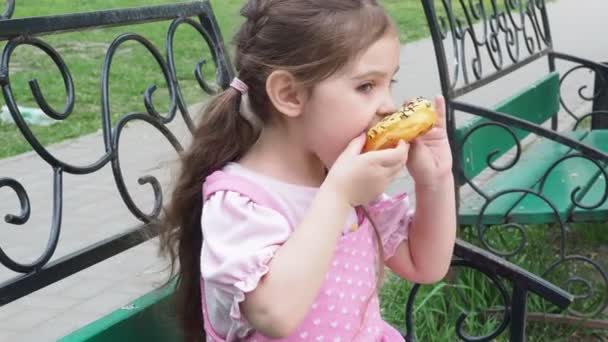 Meisje op een bankje in het park eet een donut met gele glazuur. Gelukkige jeugd, openluchtrecreatie, wandelingen. Vrolijk kind zittend op Park Bench — Stockvideo