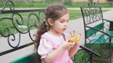 Parktaki bankta oturan kız sarı şekerli donut yiyor. Mutlu bir çocukluk, açık hava eğlencesi, yürüyüşler. Neşeli Çocuk Park Bench 'te oturuyor