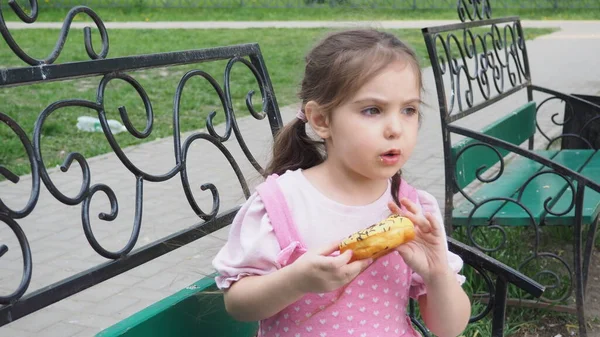 Girl on a park bench eating a donut with yellow frosting. happy childhood, outdoor recreation, walks. Cheerful Child Sitting on Park Bench — Stock Photo, Image