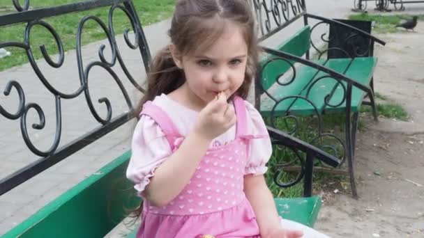 Menina em um banco de parque comendo um donut com cobertura amarela. infância feliz, recreação ao ar livre, passeios. Criança alegre sentada no banco do parque — Vídeo de Stock