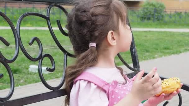 Menina em um banco de parque comendo um donut com cobertura amarela. infância feliz, recreação ao ar livre, passeios. Criança alegre sentada no banco do parque — Vídeo de Stock