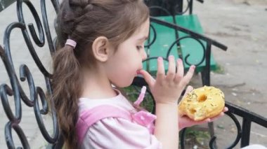 Parktaki bankta oturan kız sarı şekerli donut yiyor. Mutlu bir çocukluk, açık hava eğlencesi, yürüyüşler. Neşeli Çocuk Park Bench 'te oturuyor