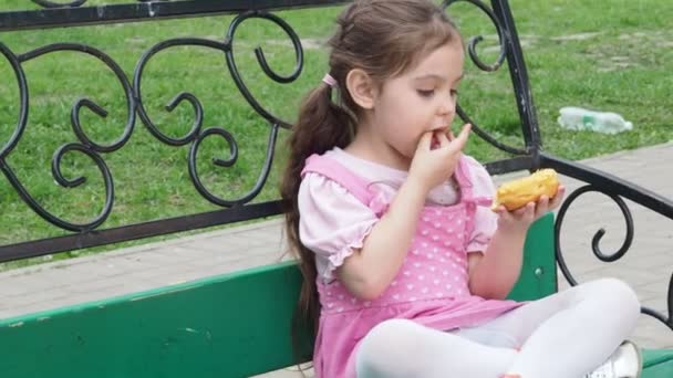 Το κορίτσι στο παγκάκι τρώει ντόνατ με κίτρινη επικάλυψη. Χαρούμενη παιδική ηλικία, υπαίθρια αναψυχή, βόλτες. Χαρούμενο παιδί κάθεται στο πάρκο Bench — Αρχείο Βίντεο