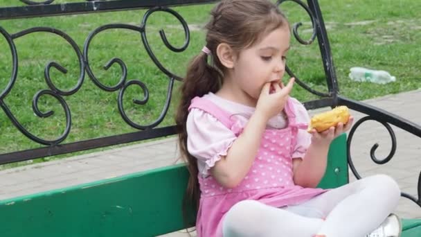 Parktaki bankta oturan kız sarı şekerli donut yiyor. Mutlu bir çocukluk, açık hava eğlencesi, yürüyüşler. Neşeli Çocuk Park Bench 'te oturuyor — Stok video