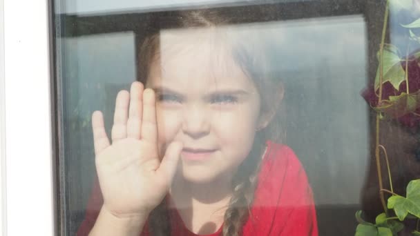 Hermosa niña sonriendo y mirando por la ventana. Un niño mira por la ventana. Retrato de niño alegre. autodistanciamiento en casa durante el virus Corona. Cuarentena Covid-19 Pandemia mundial. — Vídeo de stock