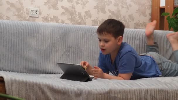 快乐的男孩躺在家里的沙发上玩Pc平板电脑。快乐的男孩玩电子游戏和大笑。在家里看卡通片或电影的青少年。在网上和朋友聊天在线学习 — 图库视频影像