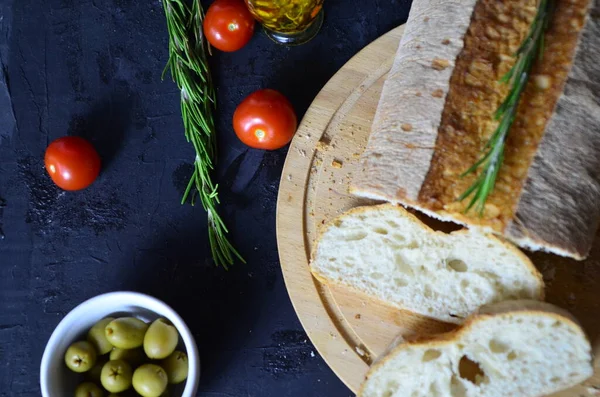 Italienisches Ciabatta-Brot in Scheiben geschnitten auf hölzernem Schneidebrett mit Kräutern, Knoblauch und Oliven vor dunklem Grunge-Hintergrund, Draufsicht — Stockfoto