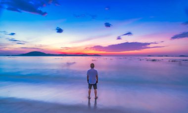 Pembe tişörtlü genç adam sahilde yürüyor.