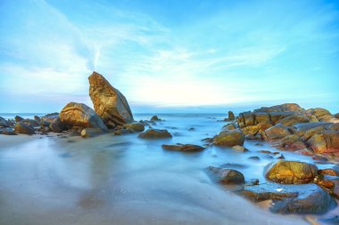 Beautiful coastal sunrise scene featuring a dramatic rock formation rising from a sandy beach.