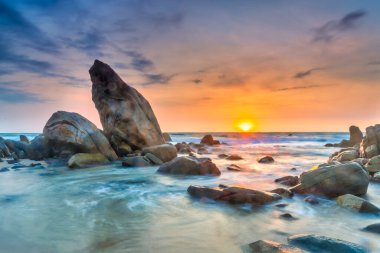 Beautiful coastal sunrise scene featuring a dramatic rock formation rising from a sandy beach.