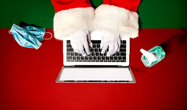 Papai Noel usando um computador portátil com uma máscara e uma garrafa de desinfetante — Fotografia de Stock