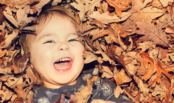 Ευχαρίστως toddler κορίτσι χαμογελά ξαπλώνοντας σε έναν σωρό των φύλλων — Φωτογραφία Αρχείου