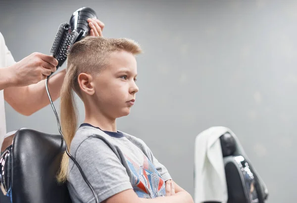 the process of hair styling a blond boy with hair dryer and comb in a chair in a barbershop salon, a barbershop concept for men and boys