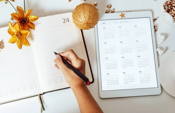 the girl writes in the diary next to the calendar for 2021 on tablet pc and wireless headphone, the concept of plans, wish list and to do list before Christmas