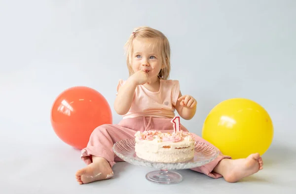 little beautiful blue-eyed girl celebrates her first birthday in the style smash a cake and she tastes her first birthday cake on a blue background with balloons