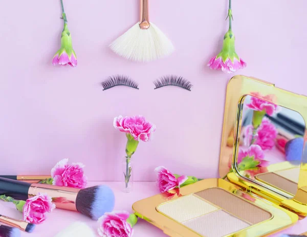 face with false eyelashes and flowers, premium makeup brushes on a colored pink background, creative cosmetics flat lay