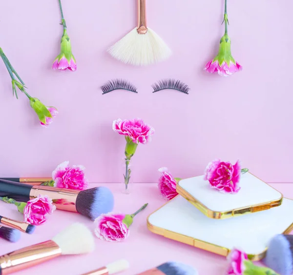 face with false eyelashes and flowers, premium makeup brushes on a colored pink background, creative cosmetics flat lay