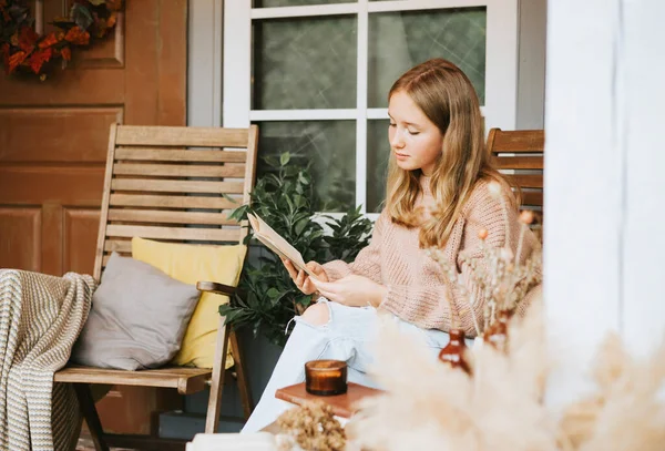 beautiful teenage girl in cozy warm knitted sweater and jeans is sitting on porch of house with cozy autumn decor and reading book, autumn season concept in pastel colors