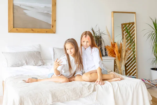 Two teenage girls in white shirts and denim shorts having fun lying on bed and using phone for selfie in a stylish modern bedroom decorated minimalistic cozy Scandinavian design, gadget addiction
