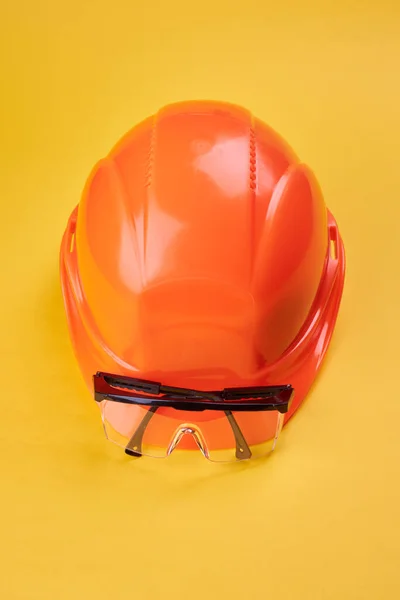 orange protective helmet and safety glasses near it on a bright yellow background. vertical orientation. protective workwear and construction industry concept