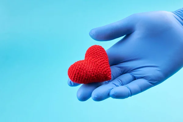 red heart symbol at persons hand with blue medical gloves on. light blue background with copy space. heart disease concept