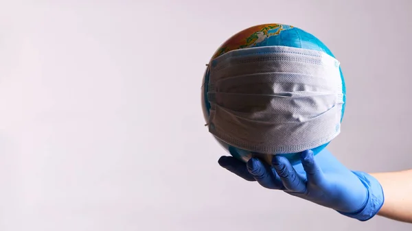 Infección epidémica global covid-19. Una persona con guantes de látex médico sostiene globo terrestre en máscara protectora del lazo del oído — Foto de Stock