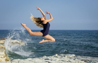 Okyanusun kıyısındaki kayalık bir sahilde dans eden genç bir kadın.