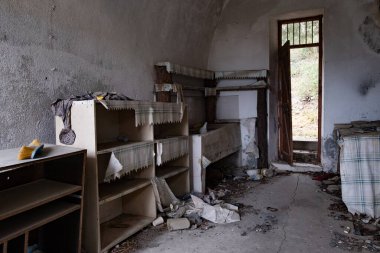 Unutulmuş bir yapının içinde, tozlu raflar ve kırık mobilyalar odayı darmadağın ediyor. Işık açık bir kapıdan süzülüyor, kalıntıları aydınlatıyor terk edilmiş ev odasını.