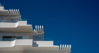 Modern beyaz bir binanın belirgin mimari özellikleri. Tasarım, parlak mavi gökyüzü ile güzel tezat oluşturan keskin çizgileri ve terasları vurguluyor..