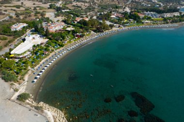 Kumlu bir sahil boyunca uzanan mavi plaj şemsiyeleri ve güneş şezlonglarıyla huzurlu kıyı şeridinin insansız hava görüntüsü. Kıbrıs Rum Kesimi 'nin tatil beldesi