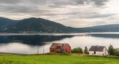 İki küçük ev, bulutlu bir gökyüzünün altında yükselen dağlarla, sakin bir göl yakınındaki yeşil bir arazide oturur. Doğa sakin ve resim gibi..