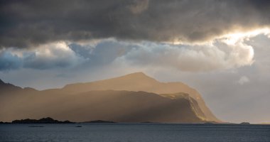 Altın güneş ışınları dramatik bulutları delip geçiyor engebeli kıyı dağlarını ve sakin suları aydınlatıyor.