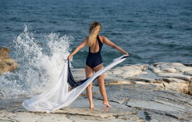 Kayalık bir sahilde, okyanusa bakan bir kadın. Arkasından dalgalar çarparken bir kumaş parçası tutuyor. Açık mavi gökyüzü ile aydınlık bir gün..