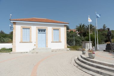 Yunan ve Kıbrıs Rum bayrakları, açık mavi gökyüzünün altında asker heykelleri ve geleneksel bir okul binası bulunan bir savaş anıtının yakınında sallanıyor. Pigenia Köyü Kıbrıs