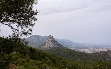 Bulutlu bir gökyüzünün altındaki engebeli bir dağ sırasının çarpıcı manzarası. Ön planda yemyeşil, uzak tepeler görkemli bir şekilde yükselirken bulutlu bir günde doğanın güzelliğini gözler önüne seriyor..