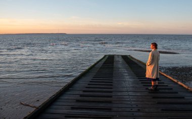 Bir iskelede tek başına duran kadın sakin denizin üzerinde gün batımını seyrediyor, Whitstable Kent, Birleşik Krallık