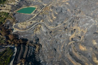 Yanmış yamaçlardan oluşan insansız hava aracı antenleri ve kararmış toprak orman yangınlarının ardından dağlık arazi boyunca uzanıyor. Yanmış ağaçlar ve toprak yollar uzaktaki hasarlı binaların yanında görülebilir..