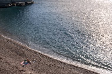 Kadın, kırmızı bir battaniyede güneşleniyor volkanik bir çakıl taşı plajında, mavi denizin yanında, parlak güneş ışığıyla.