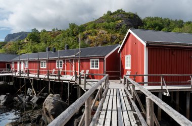 Arka planda yeşil tepeler bulunan çakıllı zeminde duran kırmızı ahşap rorbu kulübeleri Norveç kıyı mimarisini gösteriyor.