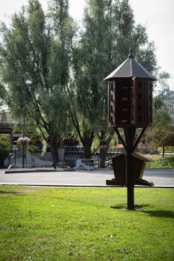 Geniş ahşap kuş yuvası, birden fazla yuva yuvası ve yeşil park ortamında bir kuş besleyicisi, barınak ve yiyecek sunuyor.