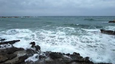 Dron 4k çözünürlüklü güçlü okyanus dalgaları bulutlu bir günde sert kayalara çarpıyor ve engebeli sahil şeridi boyunca beyaz köpük yaratıp püskürtüyor.