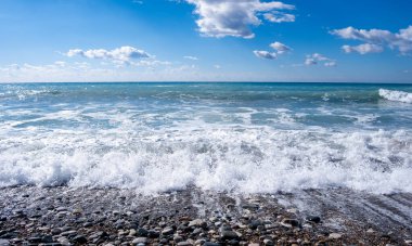 Dalgalar çakıllı bir plaja çarpıyor. Mavi bulutlu gökyüzü. Fırtınalı deniz. Okyanus dalgaları